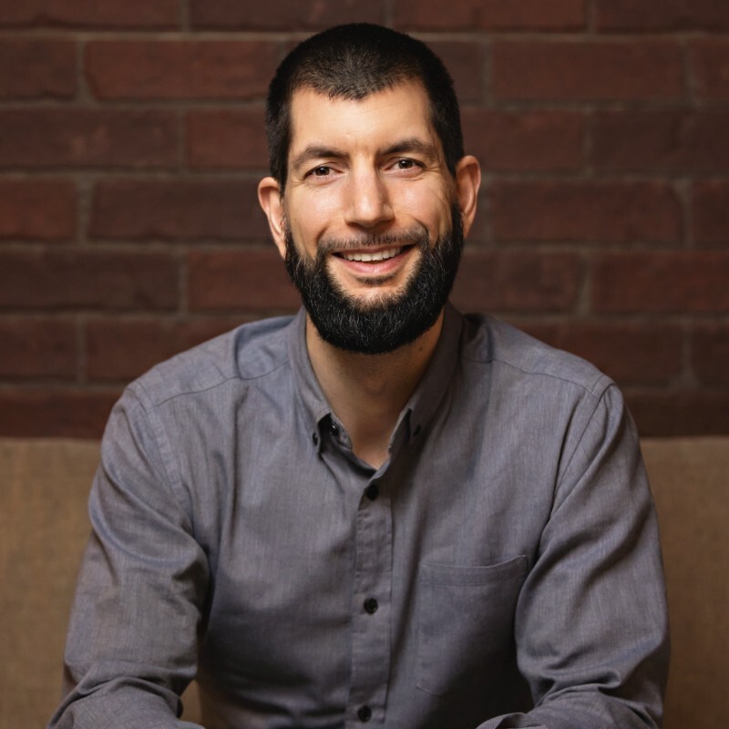 Portrait of Richard Angelo McLean smiling in a button-down shirt against a brick wall background.
